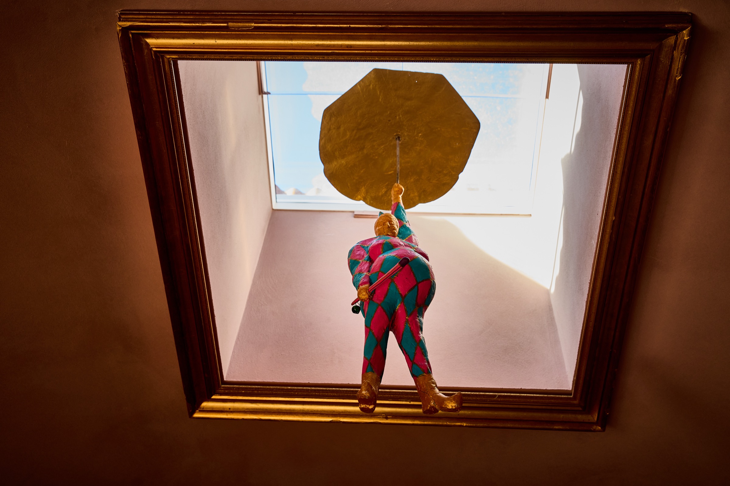 The Bell harlequin mid-flight through the skylight, gold umbrella up, seen through the gilt frame around the opening.