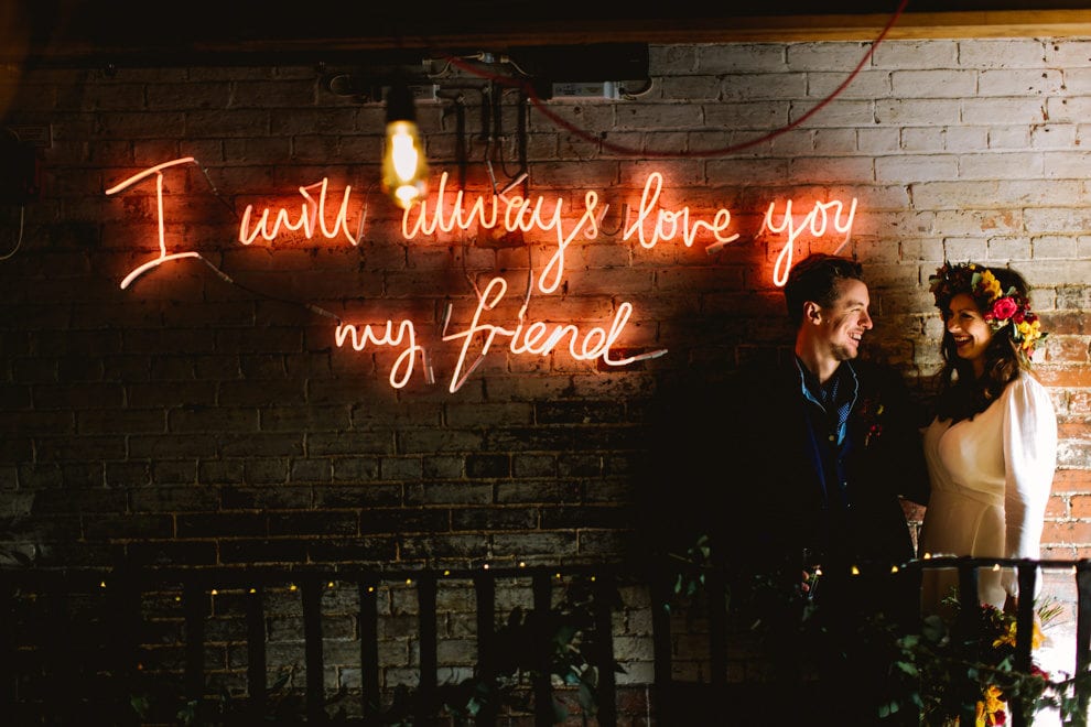 A neon sign reading I will always love you my friend, glowing on a brick wall.